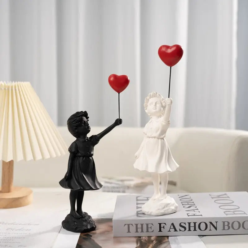 Decorative statues of a black girl and a white girl holding red heart balloons on a table with a fashion book.