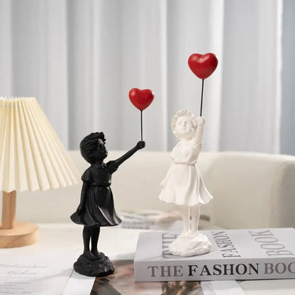 Decorative statues of a black girl and a white girl holding red heart balloons on a table with a fashion book.